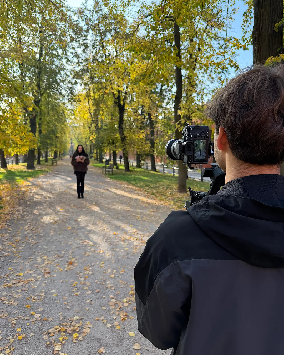 Widok zza ramienia operatora: na monitorze kamery modelka idąca jesienną aleją