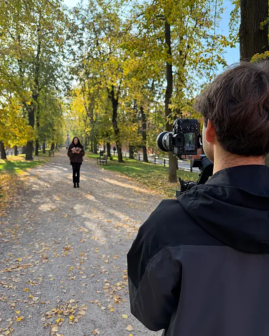 Widok zza ramienia operatora: na monitorze kamery modelka idąca jesienną aleją